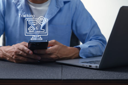 A man sits at a desk using a smartphone for tracking purposes. A laptop is placed nearby, showcasing...