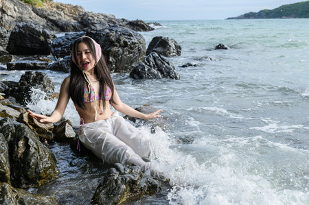 A joyful woman in a vibrant bikini sits on the rocks at the beach, splashing in the waves and...