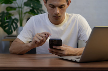 A young man is focused on his smartphone while sitting at a sleek desk with a laptop. The scene...