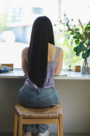 A young woman sits quietly by a window, showcasing her long hair and casual outfit. This serene...