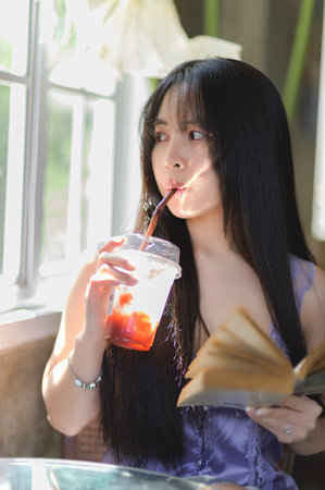 A young woman relaxes by the window, sipping a cold drink while immersed in a book, creating a serene and cozy atmosphere filled with natural light.の写真素材