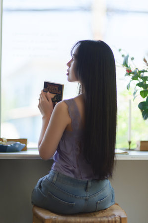 A young woman with long hair sits gracefully by a window, holding a book. Sunlight pours in,...