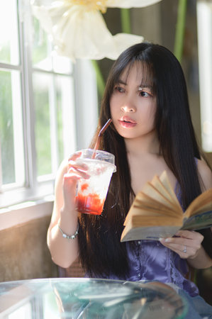 A young woman savors a refreshing drink while immersed in a book, surrounded by soft sunlight and beautiful decor, embodying tranquility and leisure.の写真素材