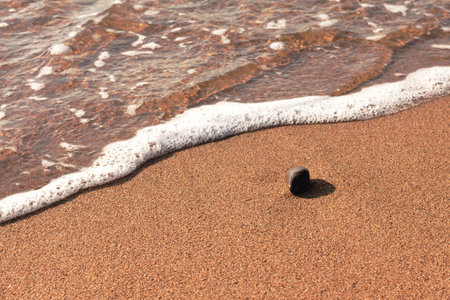 Close-up one dark brown pebble stone lies on the coast on smooth wet sand, next to the sea with clear water, fine waves and a narrow strip of white foam with bubblesの写真素材
