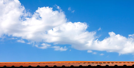 A large beautiful long white cumulus cloud in a blue clear sky over a brown metal roof