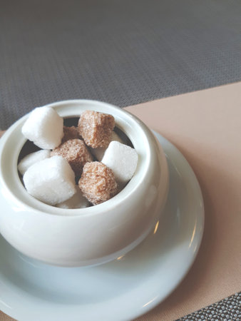 Lots of white and brown sugar cubes in a white ceramic sugar bowl on a white saucer on a table covered with a beige tablecloth and brown napkinsの写真素材