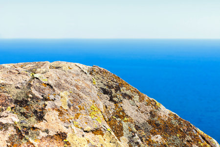 Large gray stone covered with multi-colored moss against the background of a blue calm sea and a...