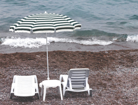 White plastic beach furniture, two sun loungers, one table and one striped beach umbrella on a...