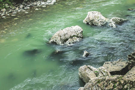 A narrow mountain river with clear green water flows between gray and white rocky banks, large gray stones in the middle of the riverの写真素材