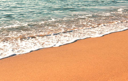 Close-up of the coastline diagonally from left to right, clean and even red-yellow sand, small waves of surf with white foam and gray-blue waterの写真素材