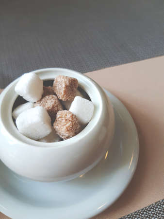 Lots of white and brown sugar cubes in a white ceramic sugar bowl on a white saucer on a table covered with a beige tablecloth and brown napkinsの写真素材