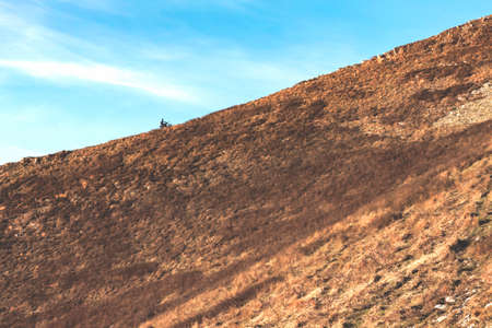 A motorcyclist on a mountain bike climbs a mountain covered with dry yellow grass, view from afarの写真素材