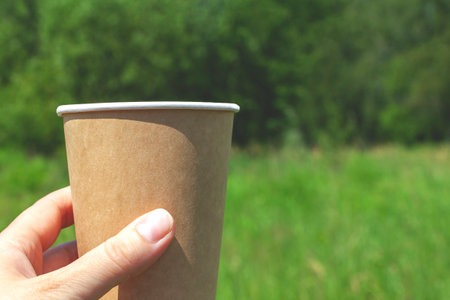 Hand holding a disposable brown cardboard cup with a drink on the left close-up on a natural background with green trees and grassの写真素材