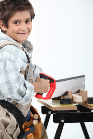 Young boy dressed as an adult carpenter cutting wood with a sawの写真素材