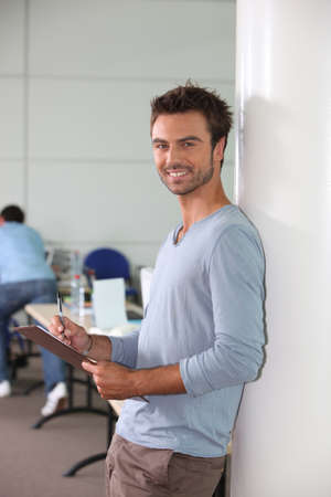 Man in office leaning against wall holding clipboard and penの写真素材