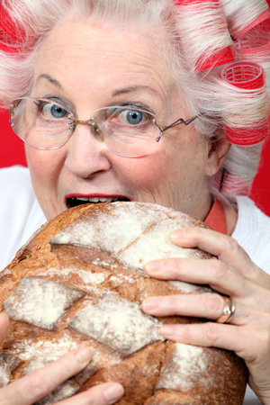 An old lady with hairroller on, biting on a loaf of bread.