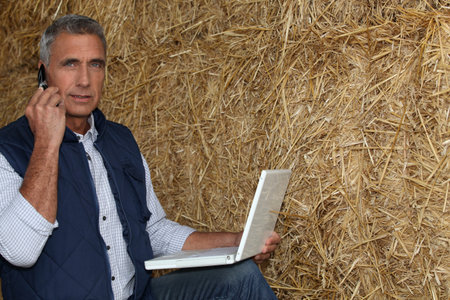mature farmer on the phone with laptop against hay background