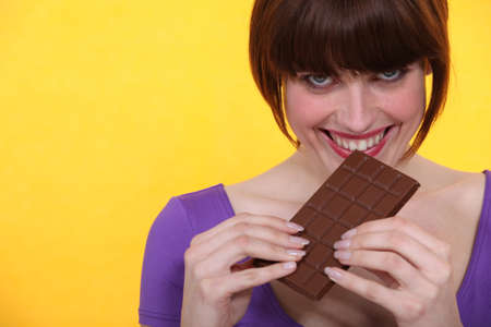 young woman relishing chocolate bar against yellow backgroundの写真素材