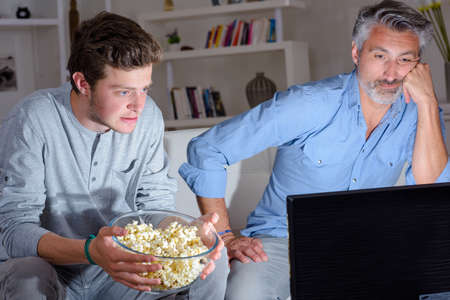 Two men watching television holding bowl of popcornの写真素材