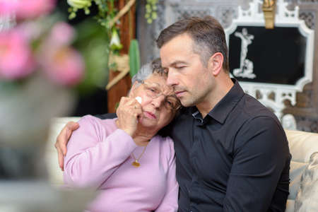 Distressed elderly lady leaning head on young man's shoulderの写真素材