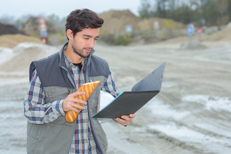 Man on building site looking at file and eating baguetteの写真素材