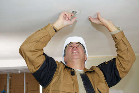 middle-age electrician fixes halogen lamp in ceiling light in roomの写真素材