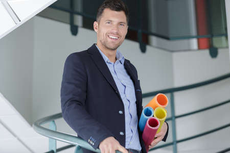 smiling male architect holding blueprints inside buildingの写真素材