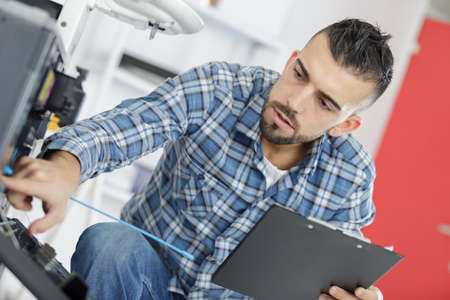 male technician sitting near dishwasher writing on clipboard in kitchenの写真素材