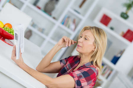 smiling woman reading book in kitchen at homeの写真素材