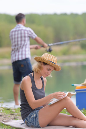 Lady sat reading a book while man fishesの写真素材