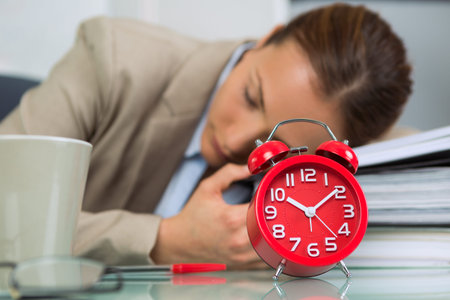 Woman asleep at office desk, alarm clock in foregroundの写真素材