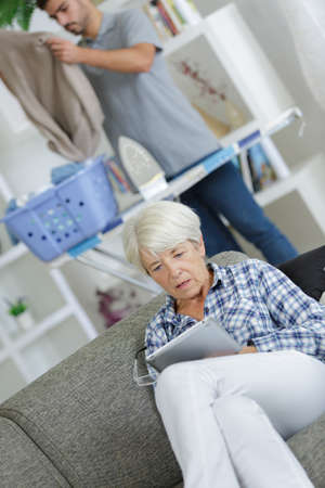 Shocked woman looking at tablet, man ironing in backgroundの写真素材