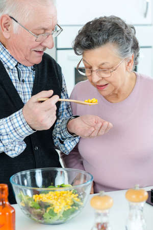 elderly man letting wife taste meal he is preparingの写真素材