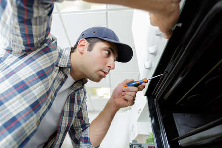 professional handyman in overalls repairing domestic oven in the kitchenの写真素材