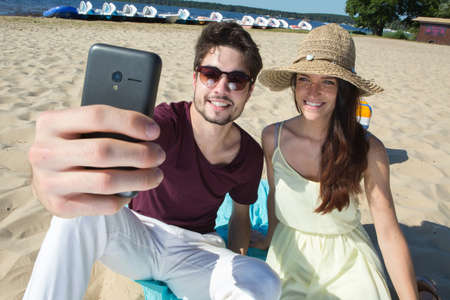 gorgeous young couple sitting at the beach and doing selfieの写真素材