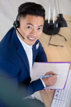 asian man wearing headset sat at computer deskの写真素材