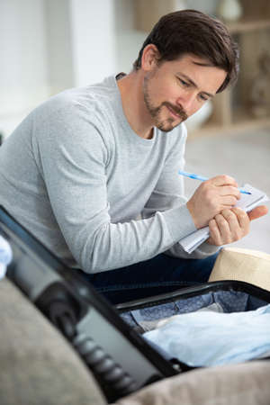 man writing list while packing his suitcaseの写真素材