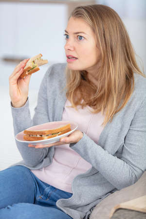 woman eating sandwich on sofa is shocked by television programの写真素材