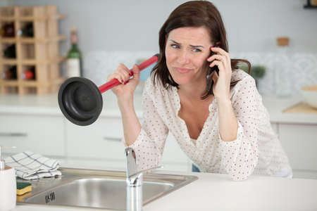woman using plunger in kitchen sink calling a plumberの写真素材