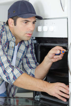 professional handyman in overalls repairing domestic oven in the kitchenの写真素材