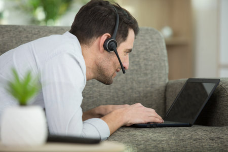 man layed on sofa wearing headset and using laptop