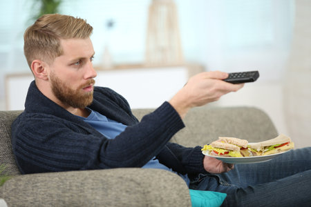 man changing tv channel with remote control while eating sandwichの写真素材