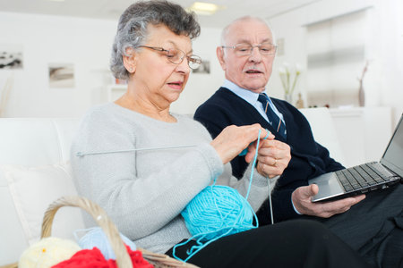 old man with computer and wife knitting sitting on sofaの写真素材