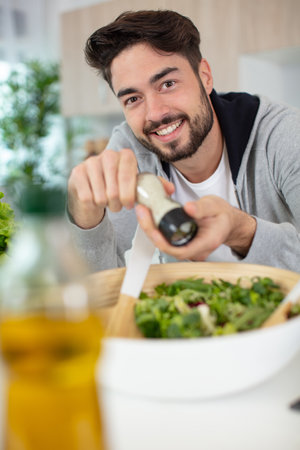 handsome smiling man salting salad in bowl at kitchen tableの写真素材