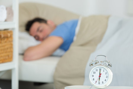alarm clock on table man sleeping in bed on backgroundの写真素材