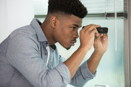 man outdoors looking through window blinds with binocularsの写真素材
