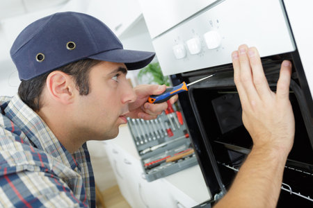 professional handyman in overalls repairing domestic oven in the kitchenの写真素材