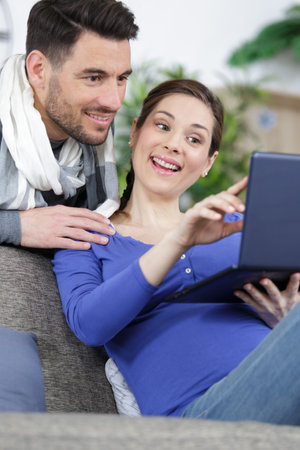 happy pregnant couple looking on laptop sat on a couchの写真素材