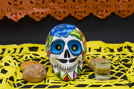 Styrofoam skull with flowers and mustache in a day of the dead offering altar with orange and yellow cut paper, bread and mezcalの写真素材