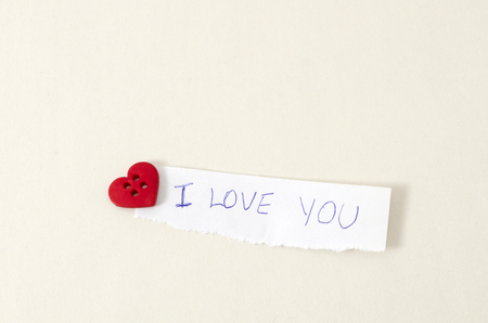Note saying I love you, written on a white piece of paper and a cute  little red heart shape button against white backgroundの写真素材